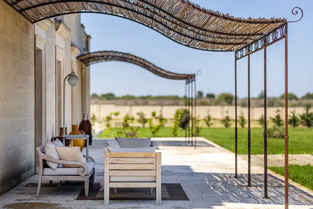 Outdoor patio with light wood furniture, cushions, and a reed pergola casting shadows on the stone floor. Green lawn and trees in the background.