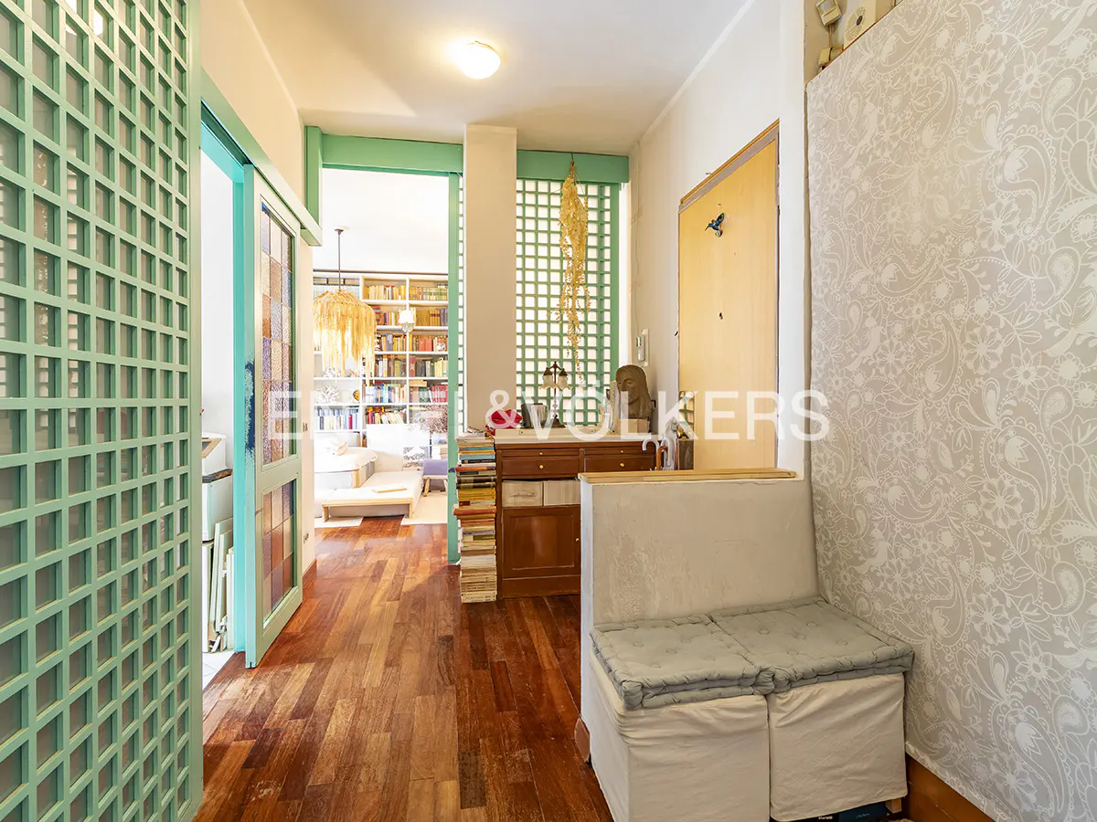 Hallway with wood floors, turquoise lattice doors, and a view into a book-filled room. A white bench sits against a patterned wallpapered wall.
