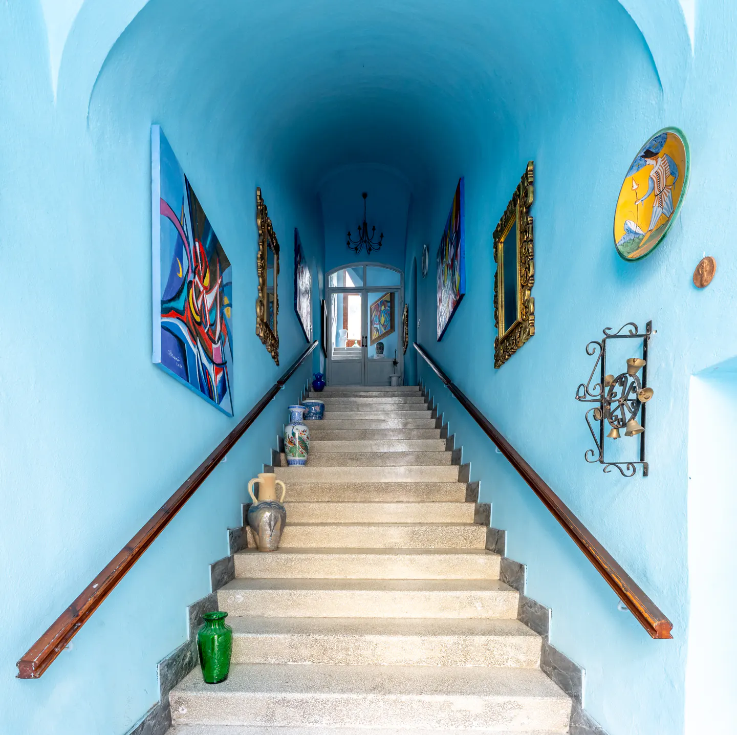 A long staircase with blue walls, decorated with art and pottery, leads to a bright doorway.