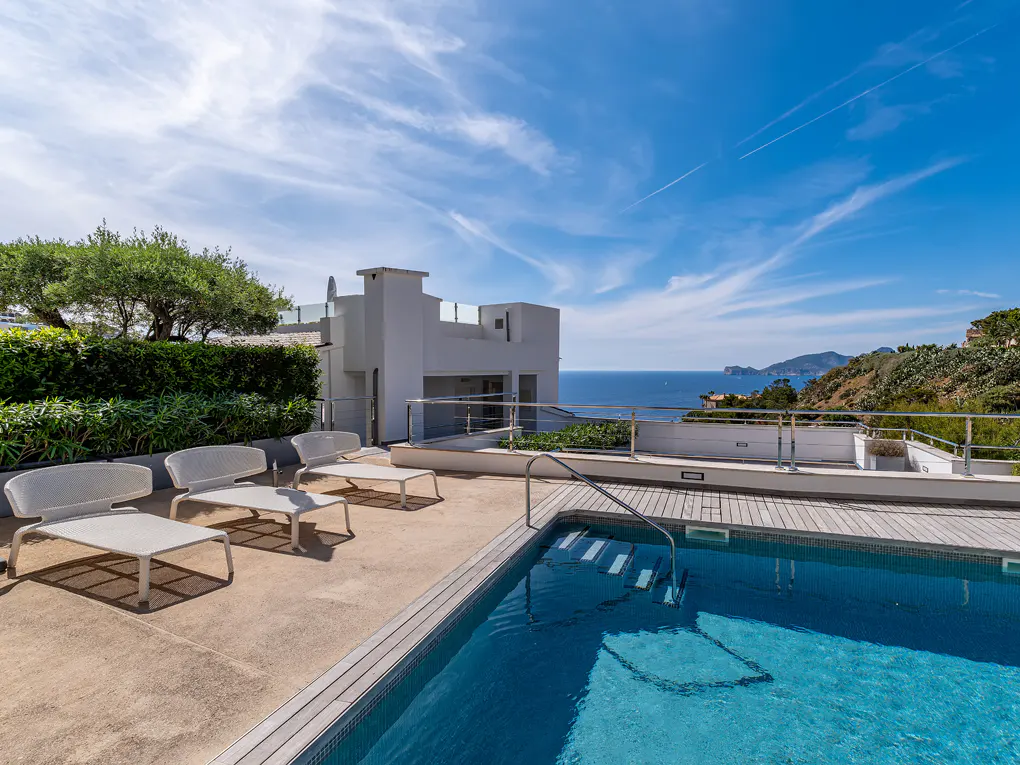 A sunny patio with a blue pool, white lounge chairs, and a modern white building overlooking the ocean.