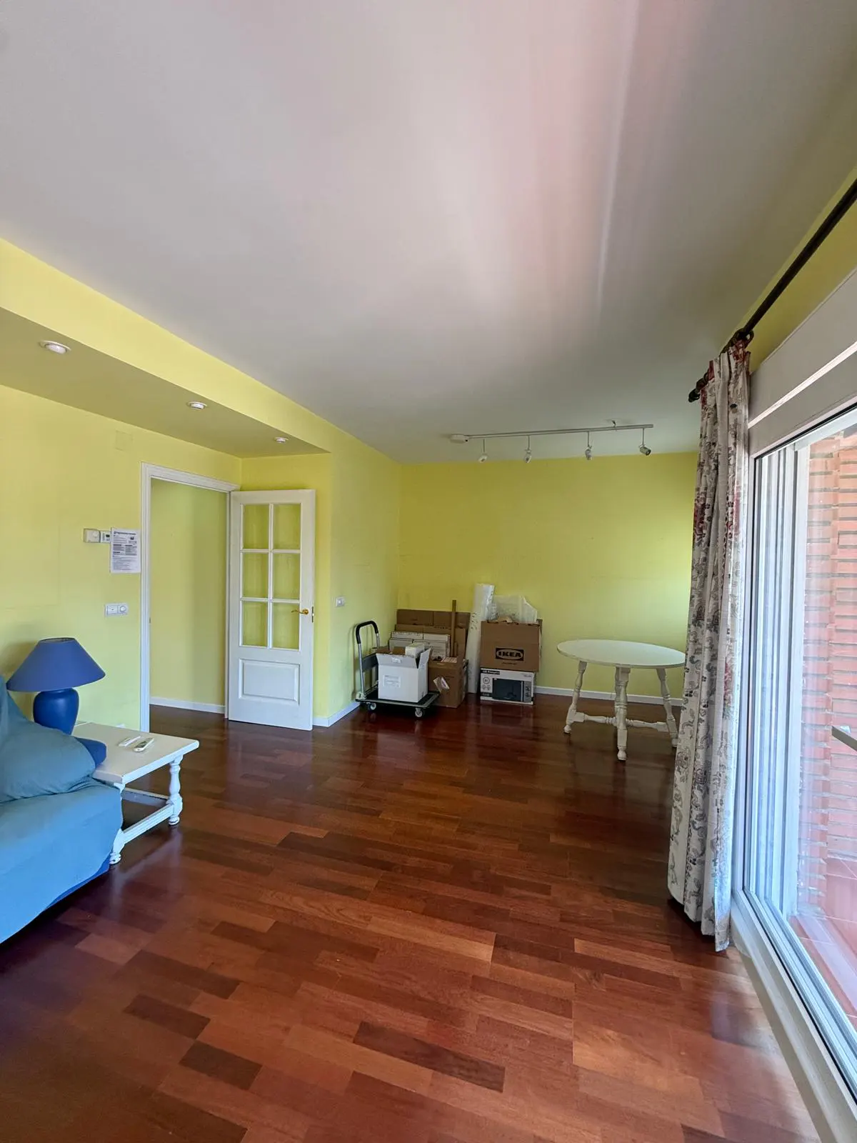 Bright, empty room with yellow walls and wood floors. A blue sofa and white table are on the left. Boxes and a white table are in the back.