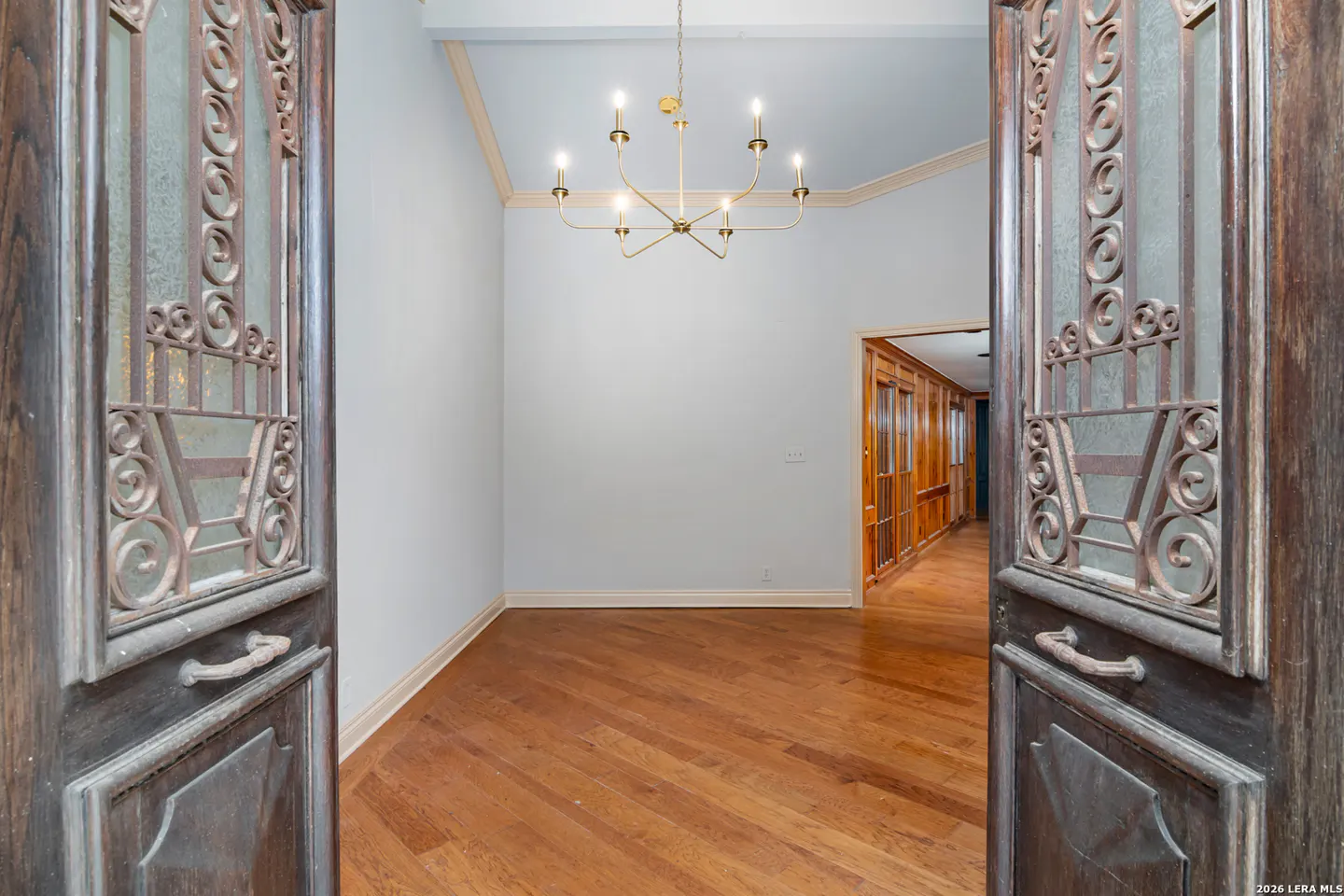View of a room with hardwood floors, light blue walls, and a gold chandelier, seen through ornate, open wooden doors.