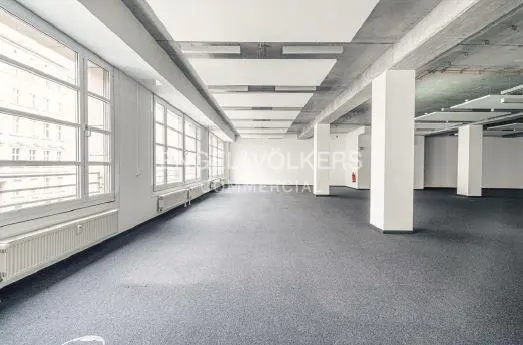 Bright, empty commercial space with gray carpet, white walls, and large windows. Concrete beams and white pillars support the ceiling.