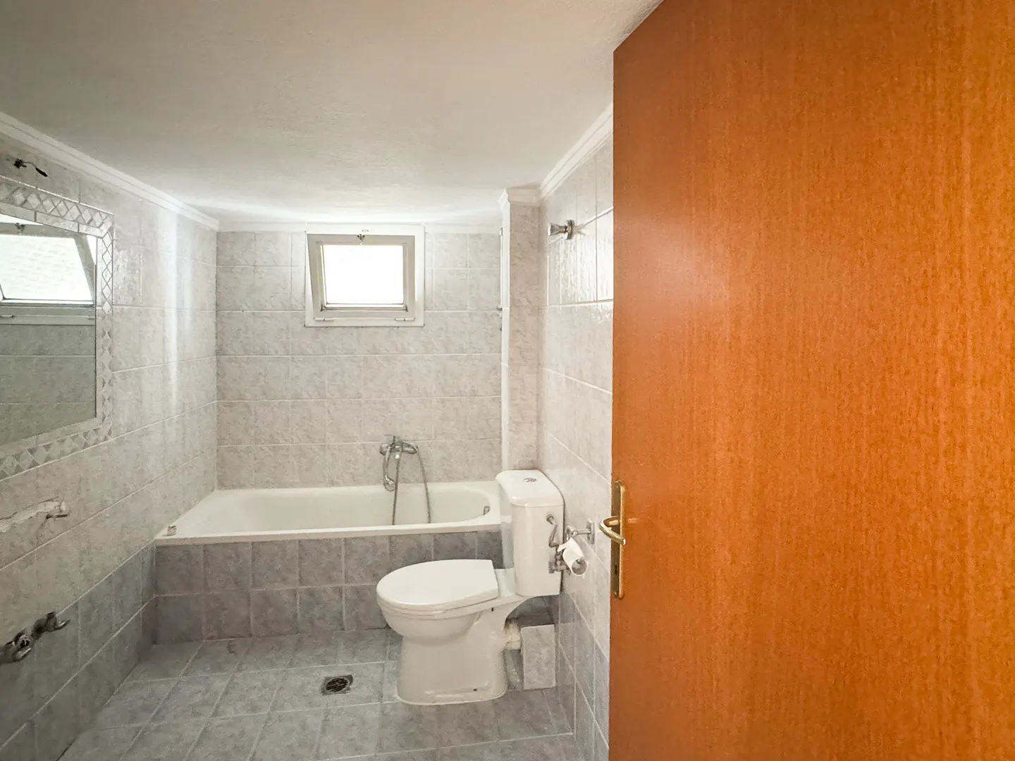 Bathroom with white toilet, tub, and gray tile. A wood door is partially open on the right. A small window is above the tub.