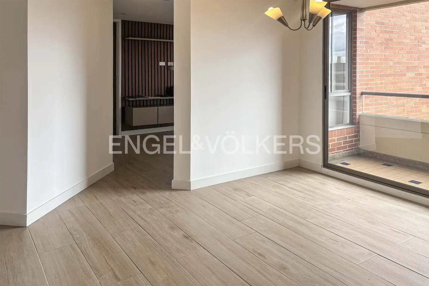 Empty room with light wood floors, white walls, and a balcony with a brick exterior. A chandelier hangs from the ceiling. Hallway visible in the background.