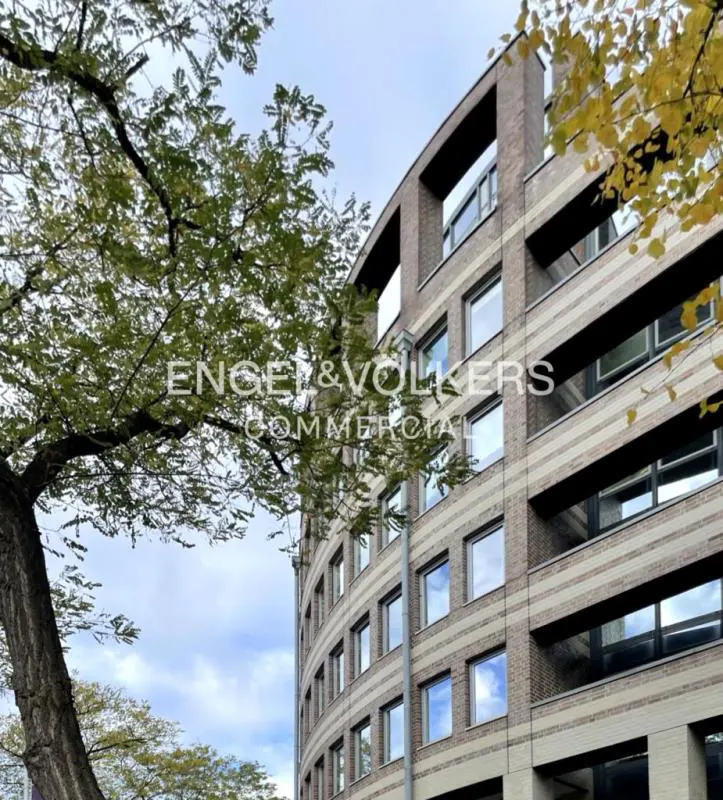 Exterior view of a curved, multi-story brick building with many windows, partially obscured by a tree. Engel & Volkers Commercial logo visible.
