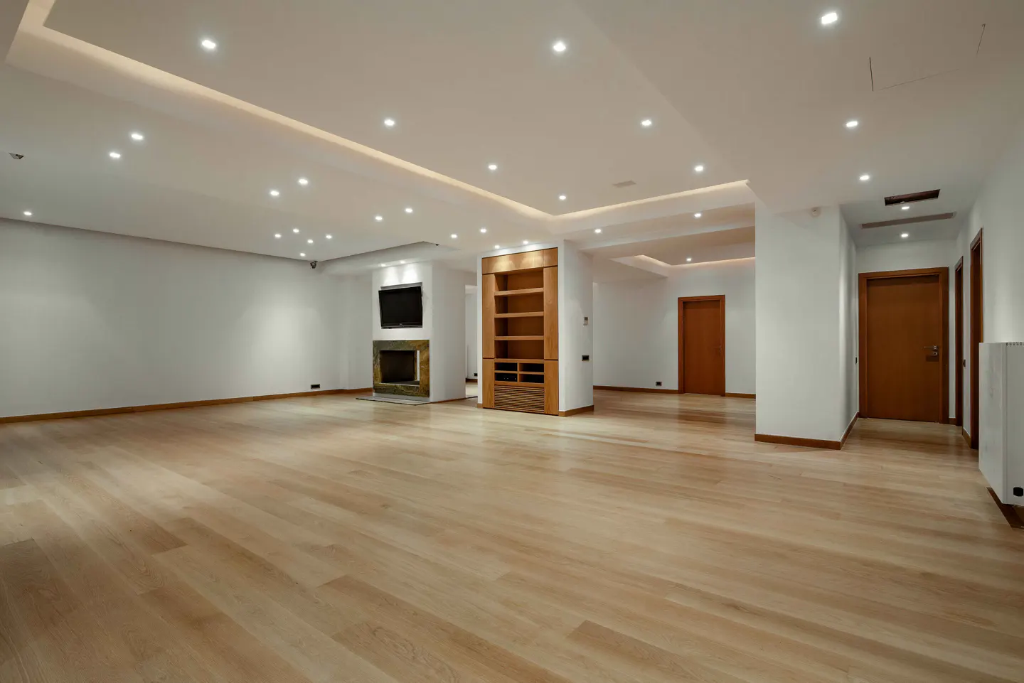 Bright, empty living room with light wood floors, white walls, recessed lighting, fireplace, TV, and built-in shelving.