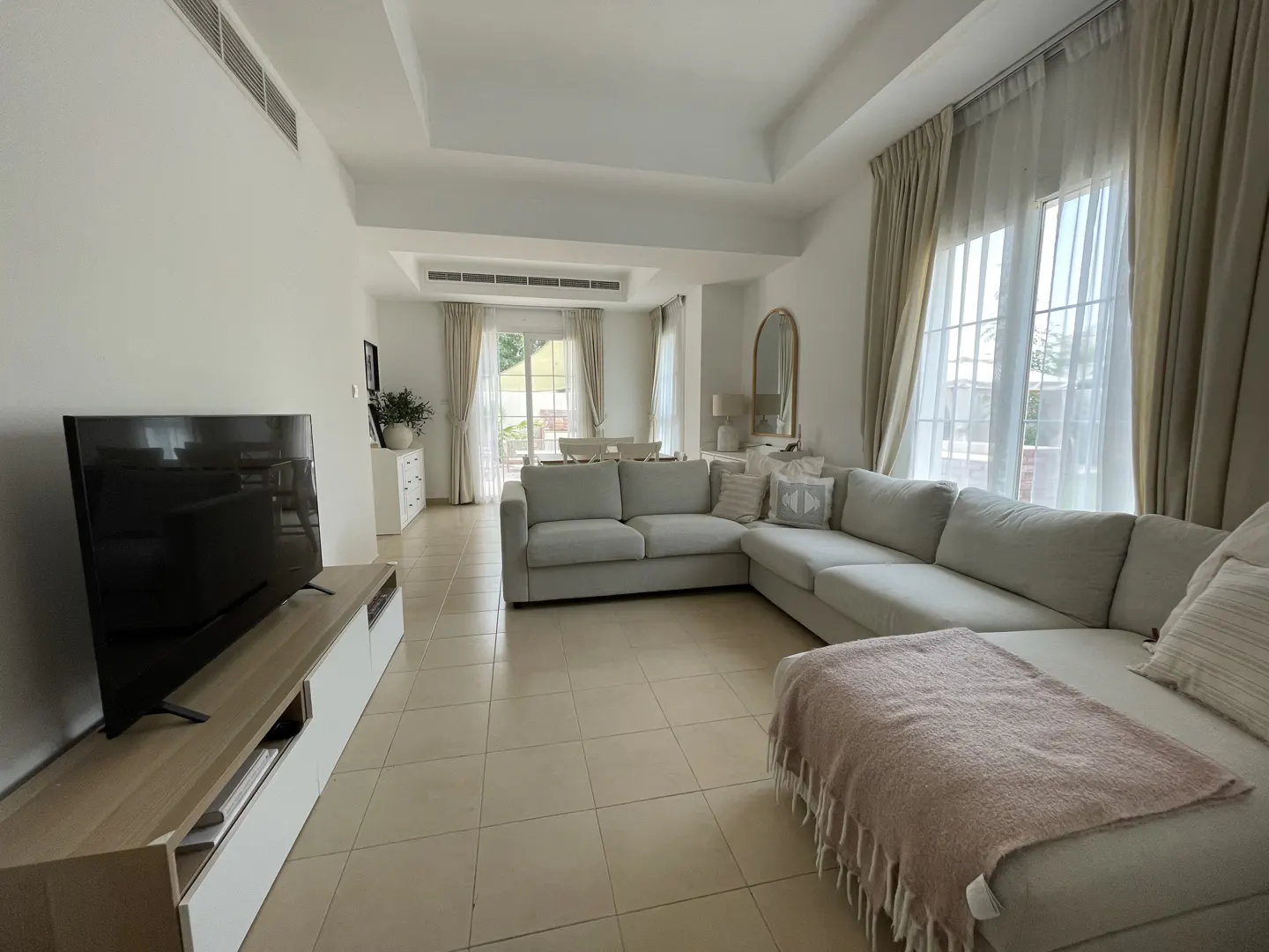 Bright living room with a large gray sectional sofa, TV on a light wood stand, and beige tile flooring. White walls and sheer curtains add to the airy feel.