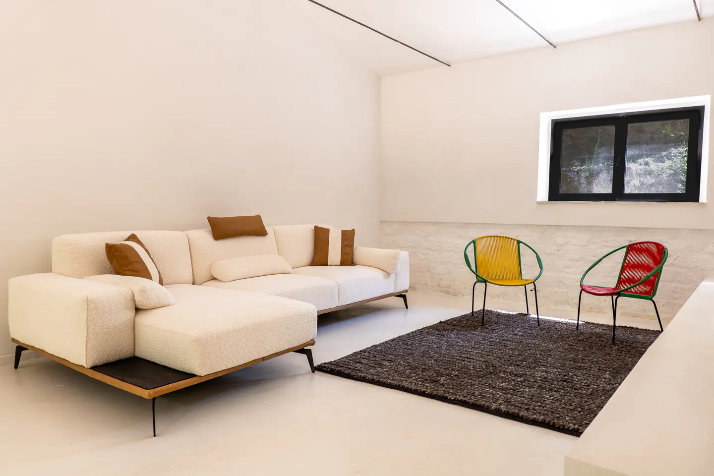 Bright living room with a white textured sofa, brown pillows, and a dark rug. Two colorful chairs sit near a window.
