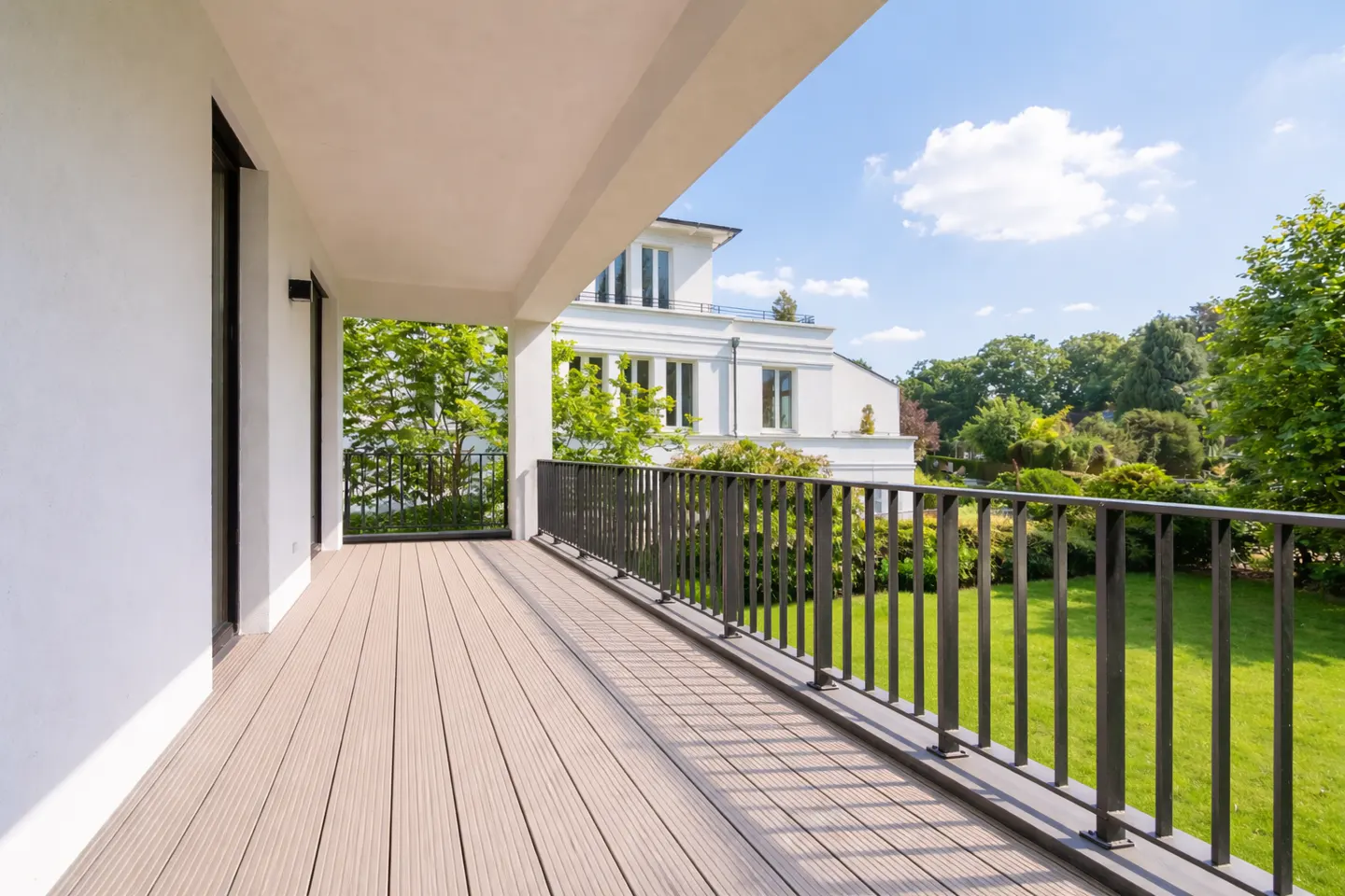 A modern balcony with a wood-look floor and black metal railing overlooks a green lawn and trees. A white building is visible in the background.