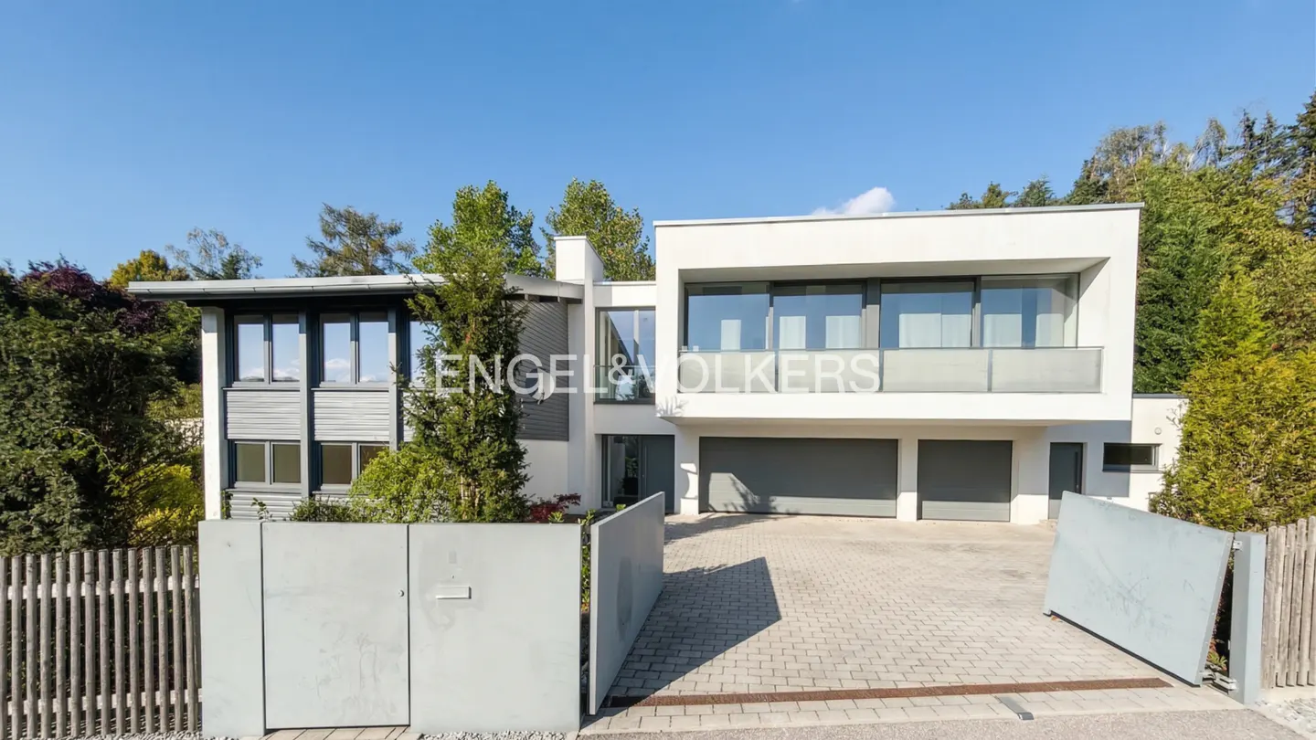 Modern two-story white house with gray garage doors and a gray gate on a brick driveway. Trees in the background.