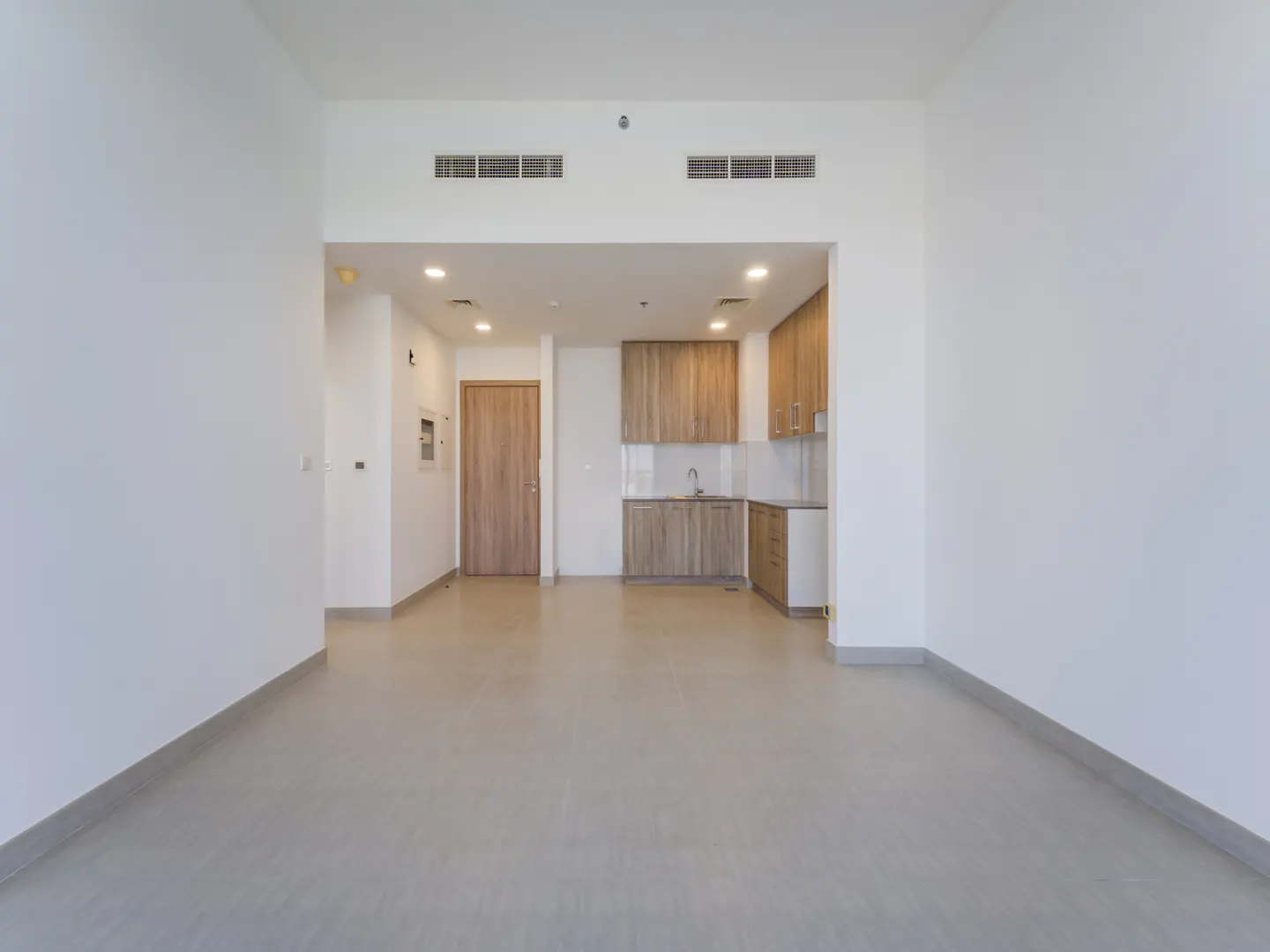 Empty apartment with white walls, gray tile floor, and a light wood kitchen in the background.