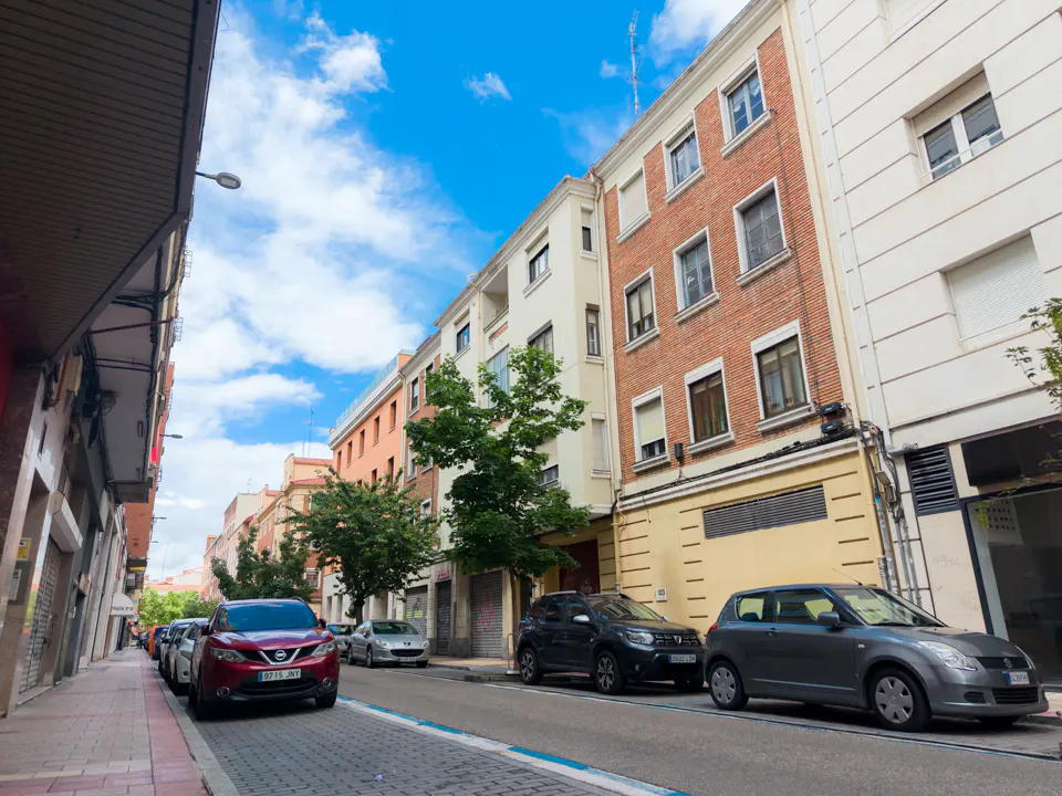 Street view of multi-story buildings with parked cars on a sunny day. Brick and white facades, blue sky with clouds.