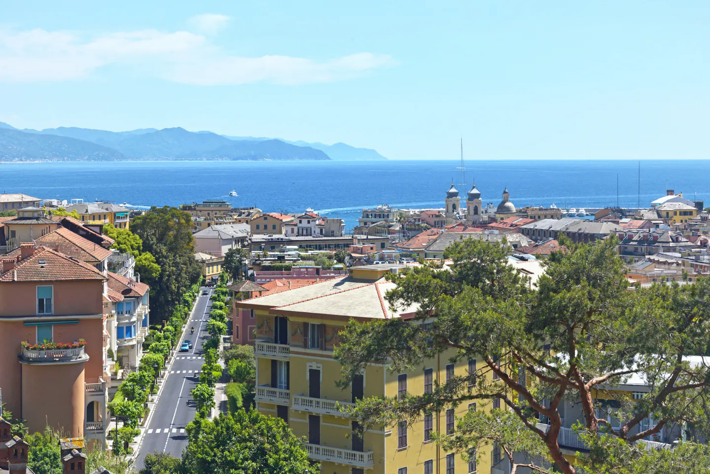 Scenic view of San Remo, Italy, featuring colorful buildings, a tree-lined street, and the blue Mediterranean Sea in the background.
