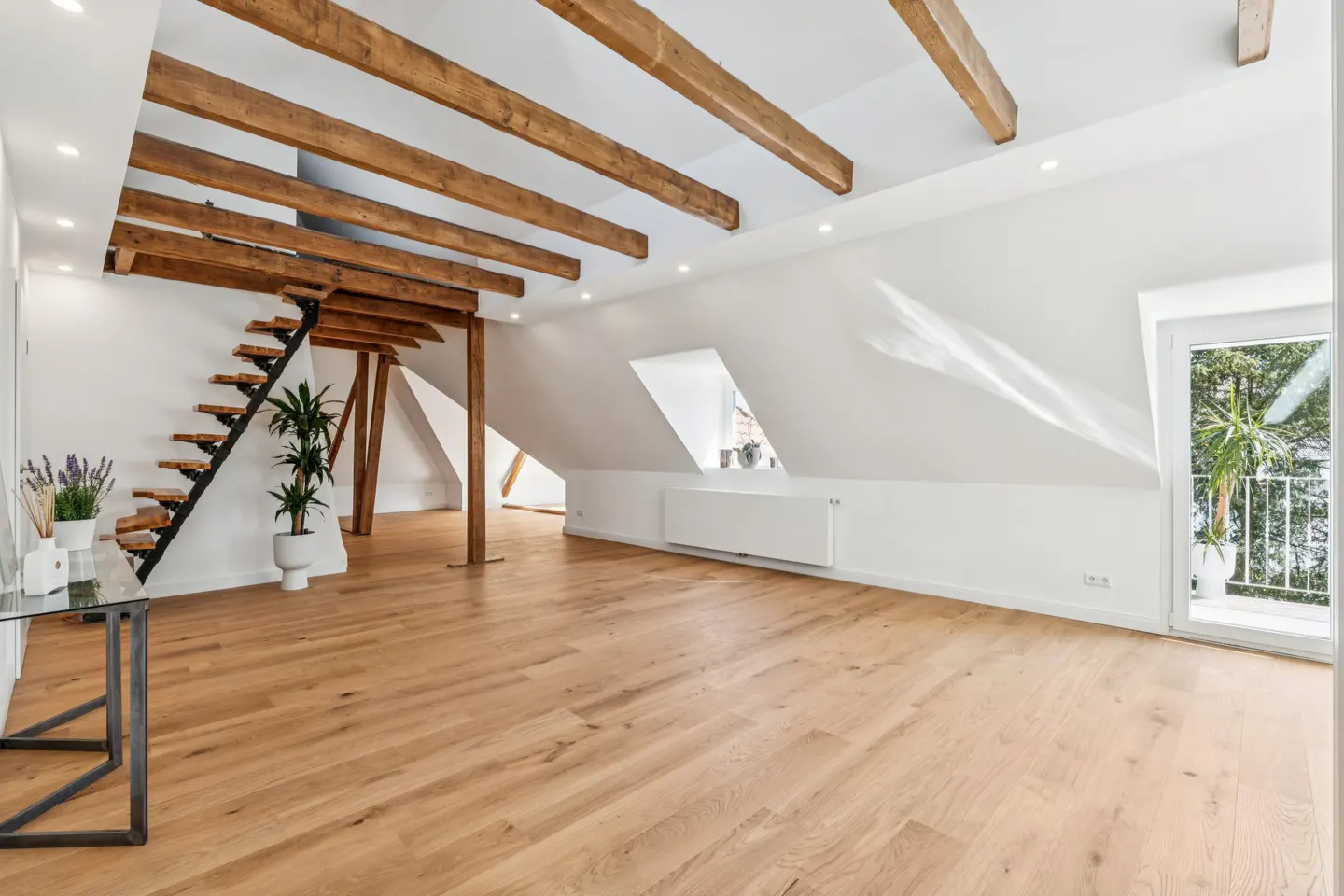 Bright attic room with wood floors, white walls, and exposed beams. Stairs lead to an upper level. Balcony with plants visible.
