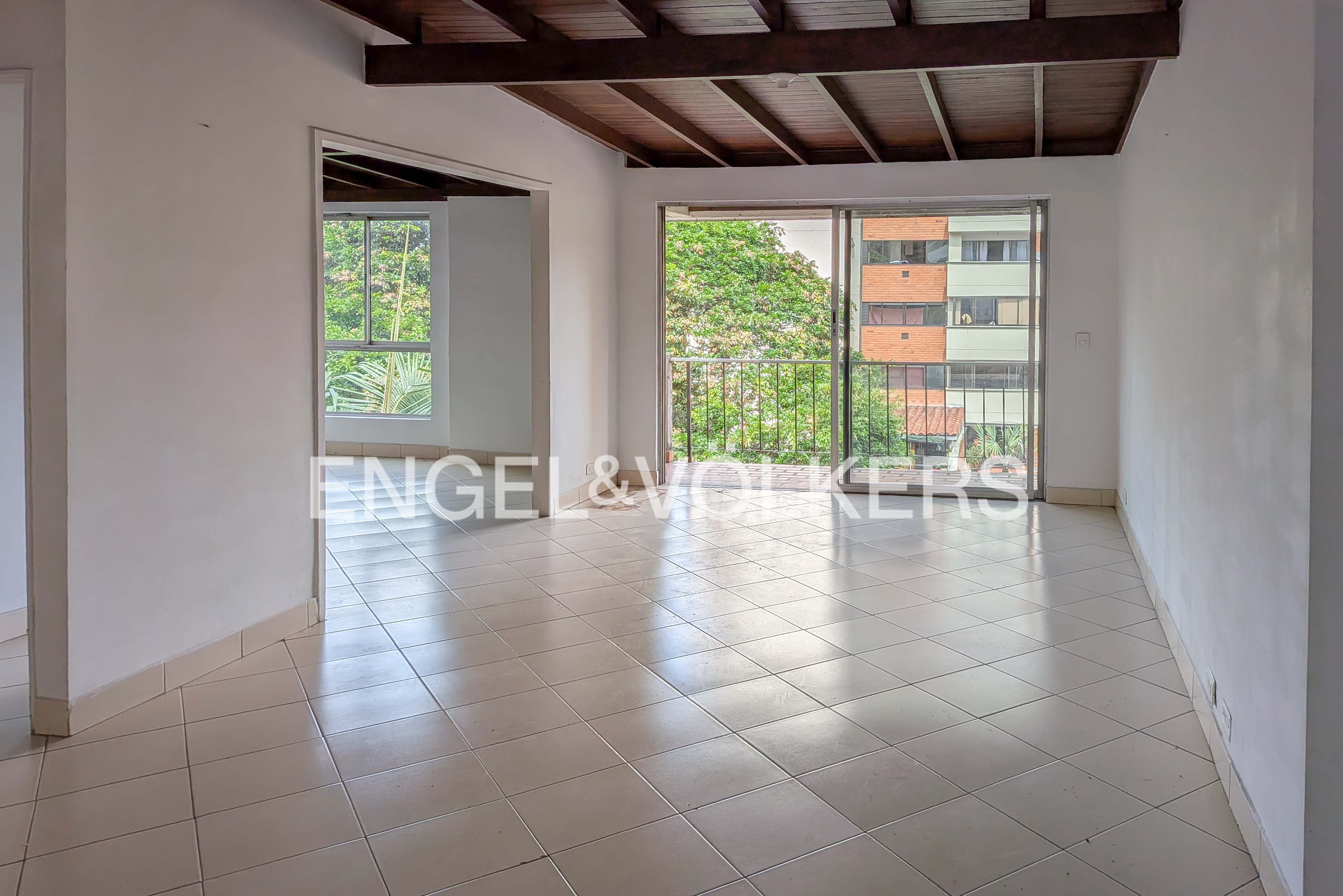 An empty room with white walls, tile floors, and a wood-beamed ceiling. A sliding glass door leads to a balcony with a view of trees and a building.