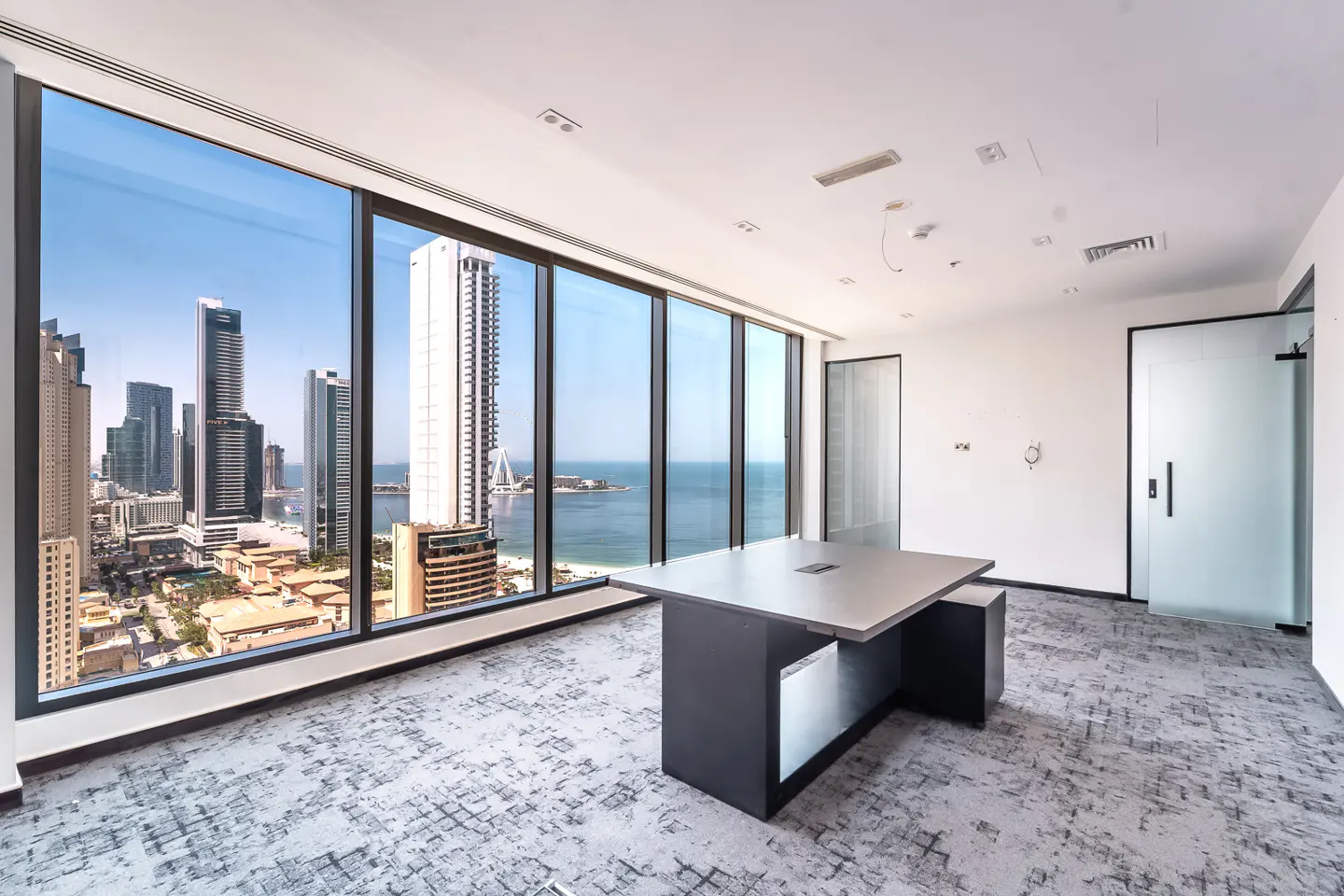 Bright, empty office space with a large window view of a city skyline and ocean. A gray desk sits on a patterned gray carpet. White walls and a frosted glass door are visible.
