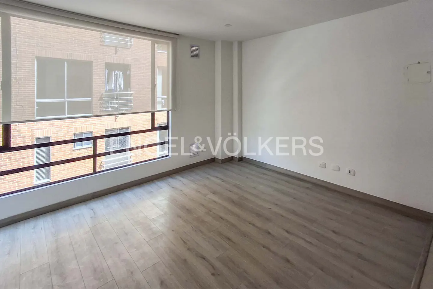 Empty room with light wood floors, white walls, and a large window overlooking a brick building.