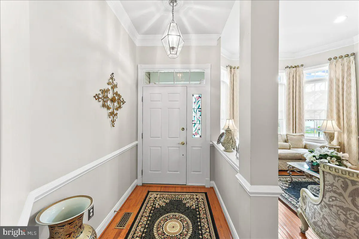 A bright foyer with a white front door, hardwood floors, and a patterned rug. A living room is visible in the background.
