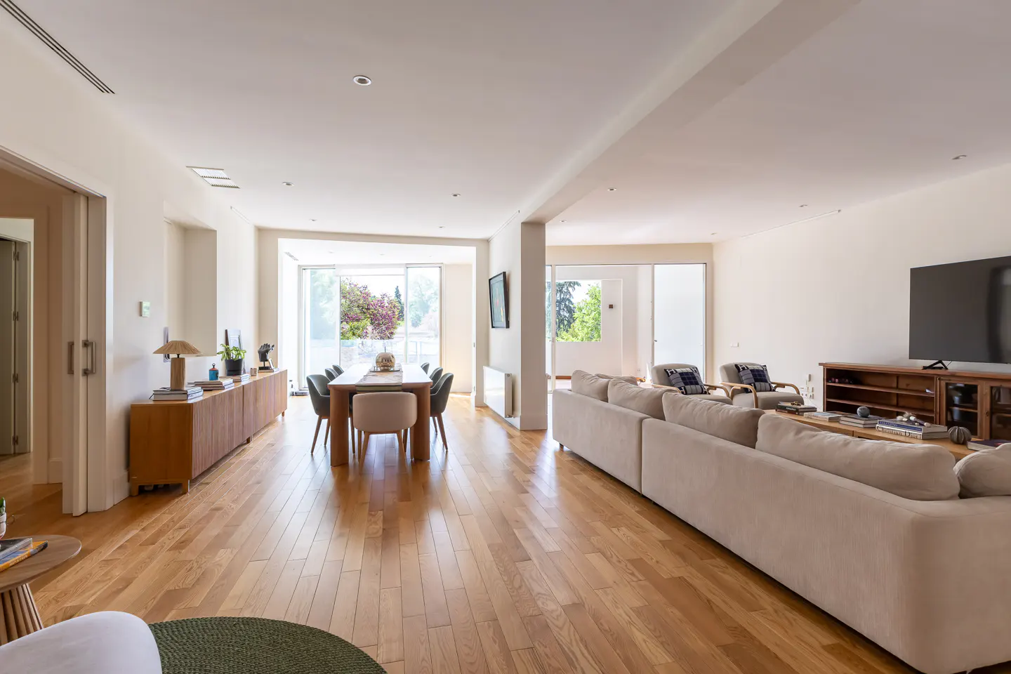 Open-concept living space with hardwood floors, a dining table, a beige sofa, and a large TV. Natural light floods the room.