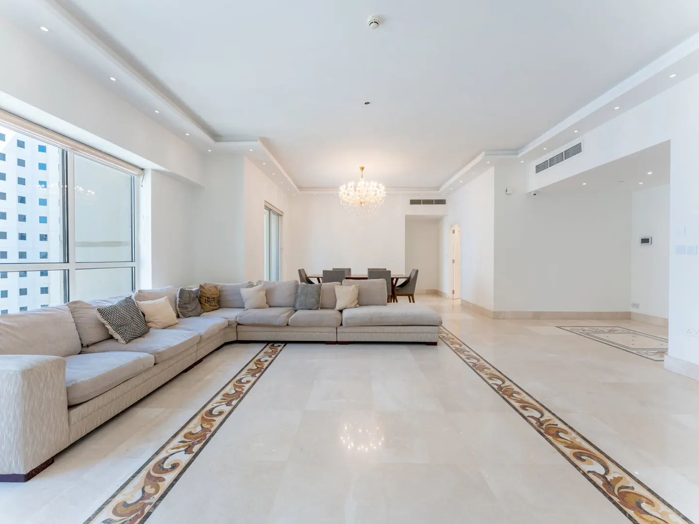 Bright, open living room with a large beige sectional sofa, marble floors with decorative borders, and a dining table in the background.