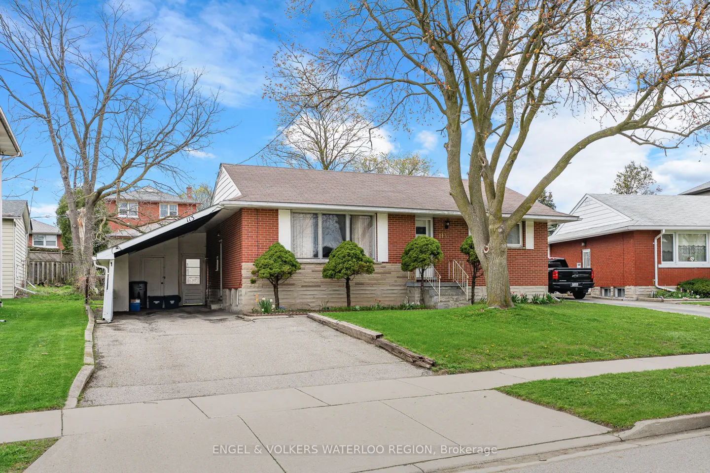 A red brick bungalow with a brown roof, a carport, and a green lawn on a sunny day.