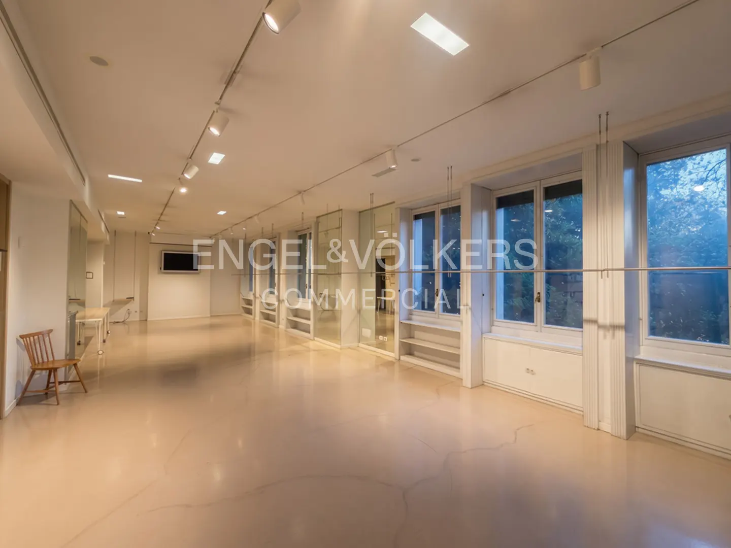 Spacious, empty commercial office with white walls, polished light-colored floors, track lighting, and large windows.