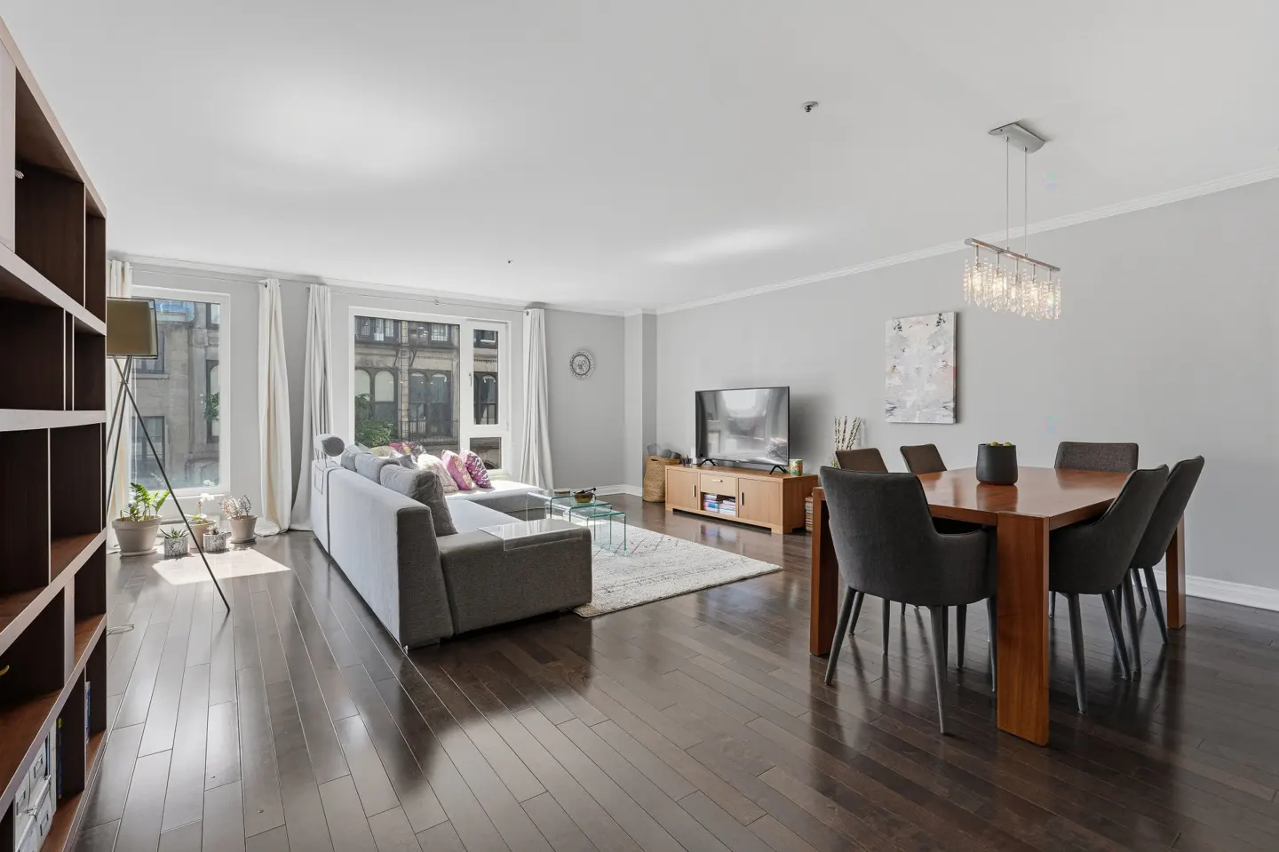 Bright open-concept living and dining room with dark hardwood floors, a grey sectional sofa, and a wooden dining table.