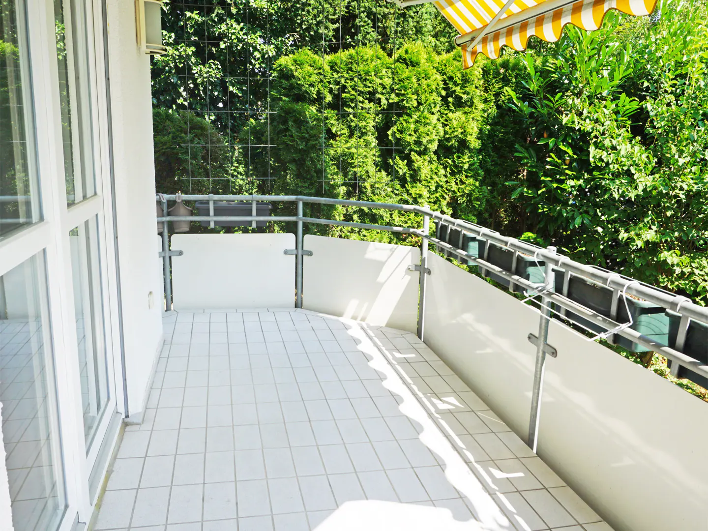 A bright balcony with white tile flooring, a metal railing with white panels, and green trees in the background.