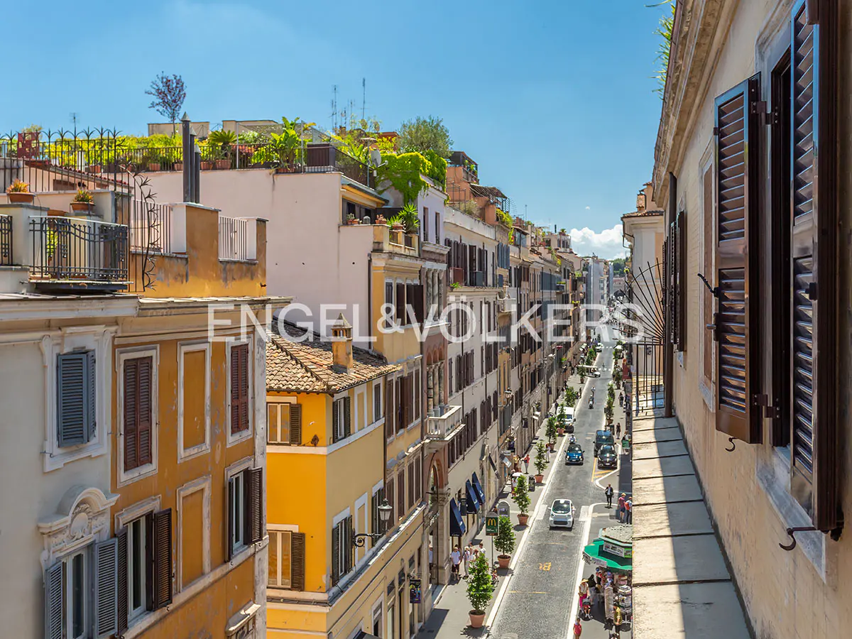 View from a balcony overlooking a narrow, sunlit street in Rome lined with historic yellow and beige buildings.
