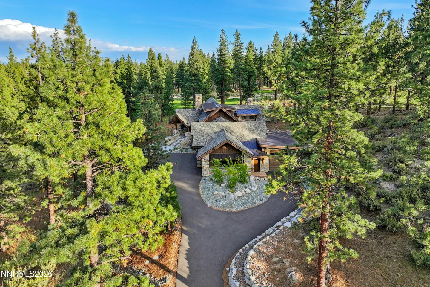 Aerial view of a stone and wood home with a shingled roof, surrounded by tall green trees and a blue sky.