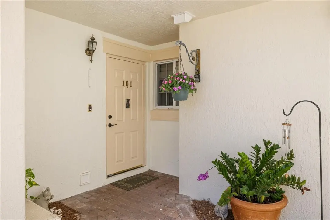 Exterior view of unit 101 with a beige door, brick walkway, and potted plants. A hanging flower basket adds color.