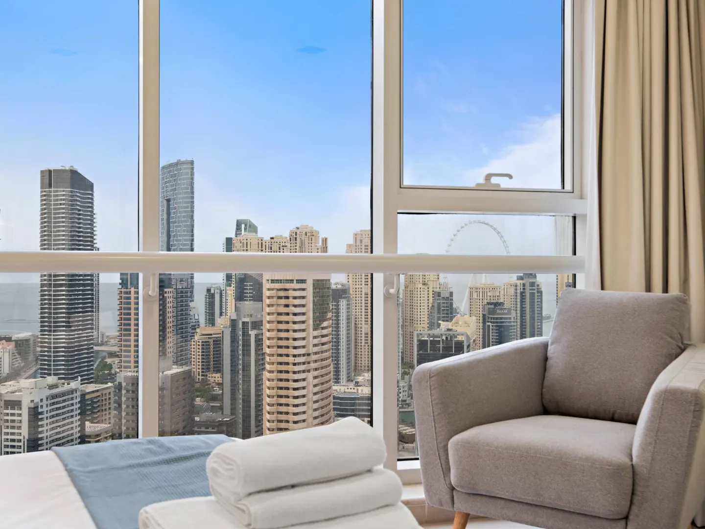 Condo interior with a gray armchair, white towels on a bed, and a large window overlooking a city skyline with skyscrapers and a Ferris wheel.