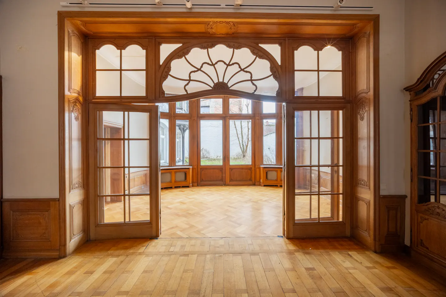 Ornate wooden doorway with glass panes leads to a sunroom with herringbone wood floors.