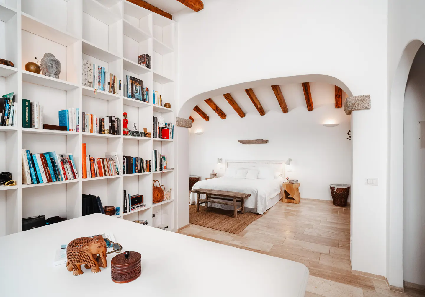 Bright bedroom with white walls, arched doorway, and wooden beams. A large bookshelf is filled with books and decorative objects.