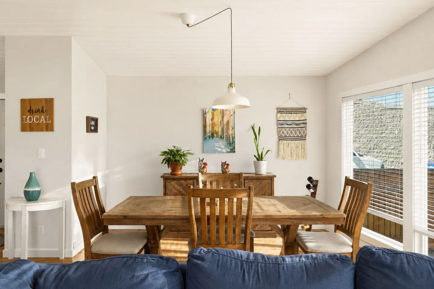 Dining room with a wooden table, chairs, a sideboard, wall art, and a pendant light, viewed from behind a blue sofa.