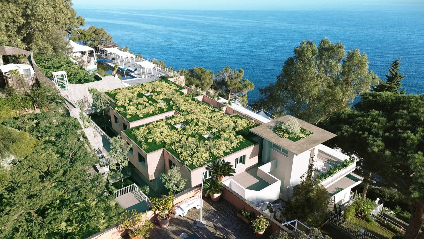 Aerial view of a pink building with green roofs, overlooking the blue ocean. Lush greenery surrounds the property.