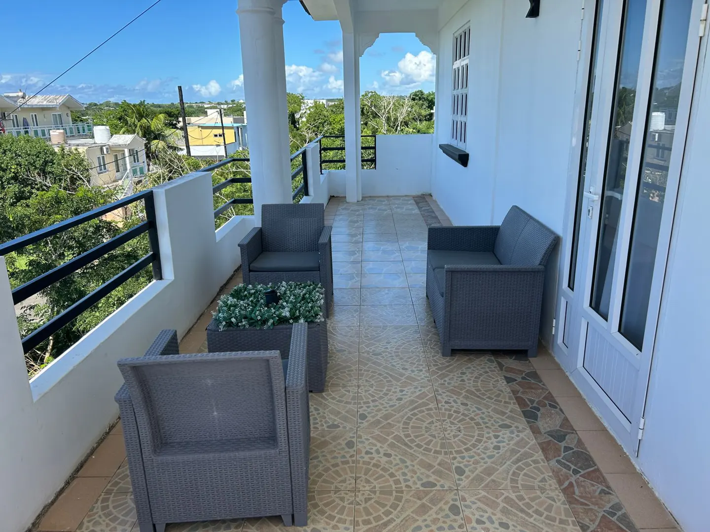 Outdoor patio with gray wicker furniture, including chairs and a loveseat, on patterned tile. Black railing and white columns frame a view of trees and buildings.