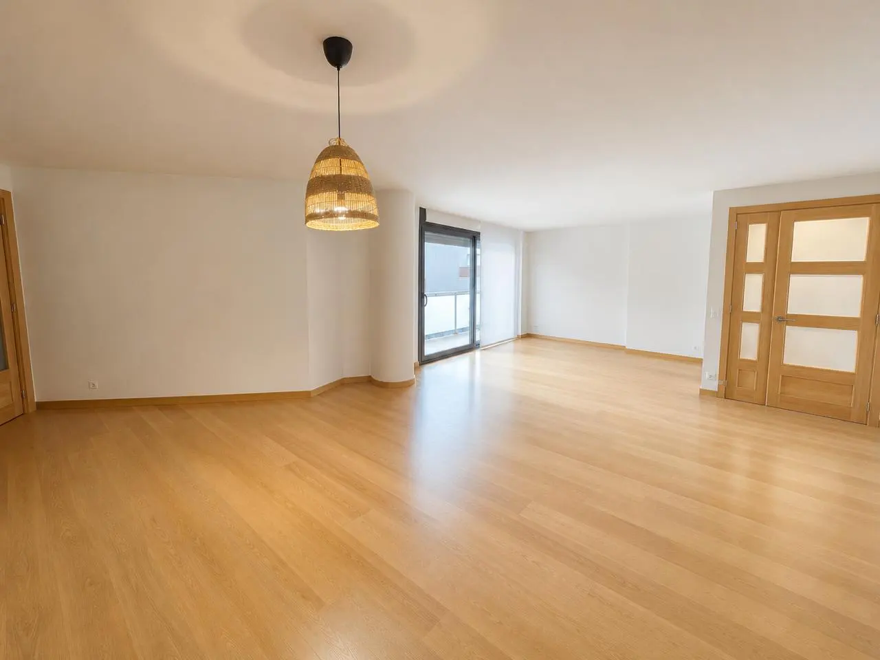 Empty room with light wood floors, white walls, and a woven pendant light. Sliding glass doors lead to a balcony.