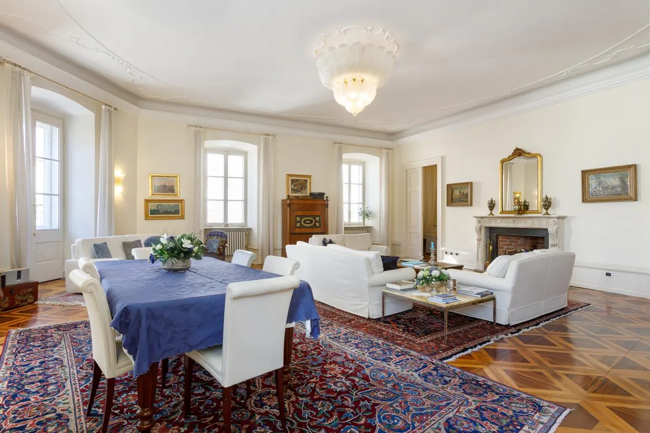 Bright, spacious living room with white sofas, a fireplace, and a dining table with a blue tablecloth. A patterned rug covers the wood floor.