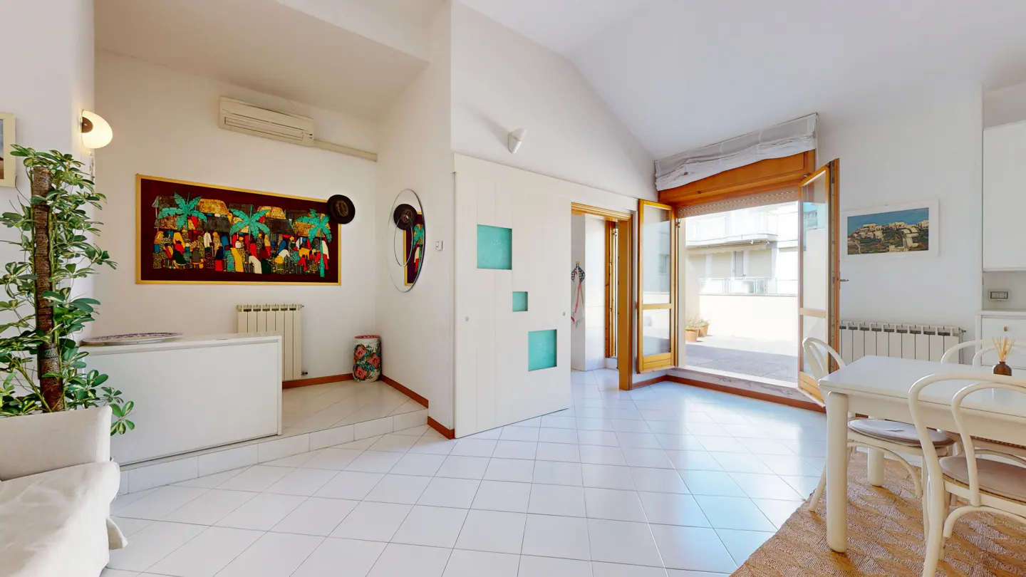 Bright, airy room with white tile floors, a dining table, and an open door to a patio. Artwork and plants add color.
