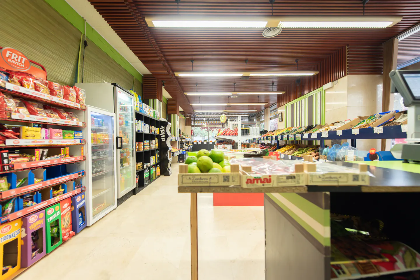 Interior view of a grocery store with shelves of snacks, drinks, and produce under a brown slat ceiling with fluorescent lights.