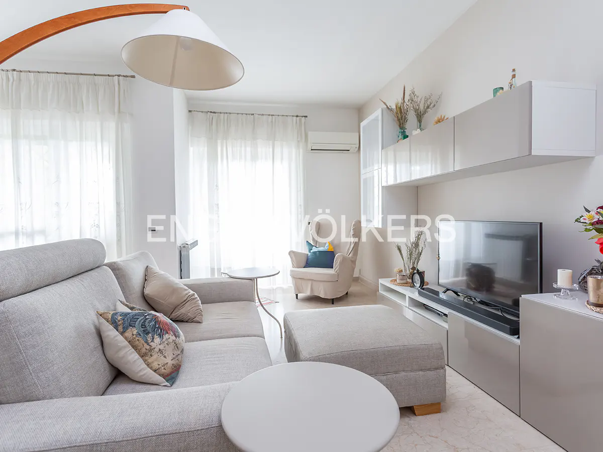 Bright living room with a gray sofa, ottoman, white cabinets, and a curved floor lamp. Natural light filters through sheer curtains.