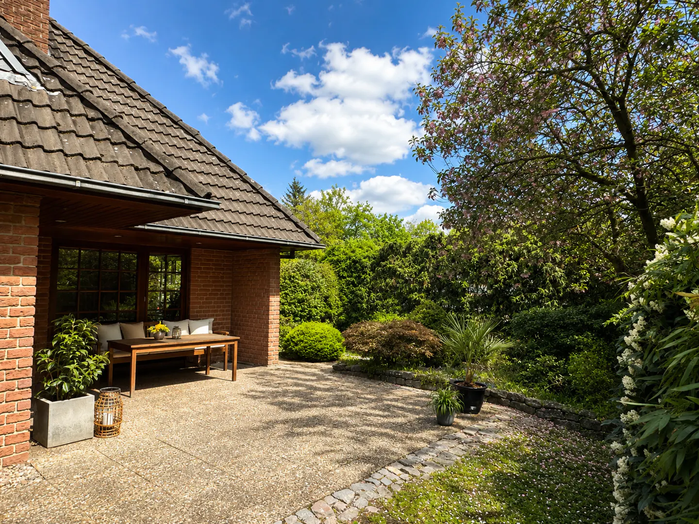 Brick house with a patio, table, and lush green garden under a blue sky with white clouds.