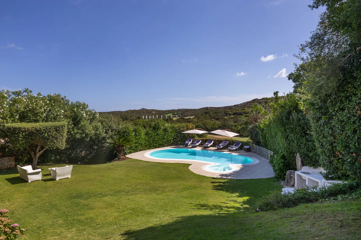 A backyard with a pool, lounge chairs, and white chairs on a green lawn under a blue sky.