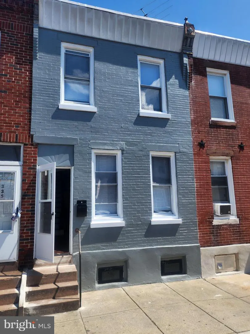 Two-story row house with gray brick, white trim, and an open white front door. A red brick house is on the right.
