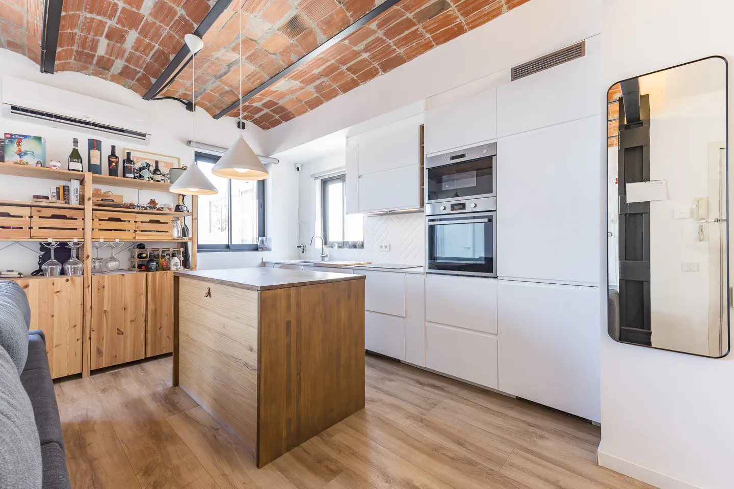 Modern kitchen with a brick vaulted ceiling, wooden island, white cabinets, stainless steel oven, and a wall mirror.
