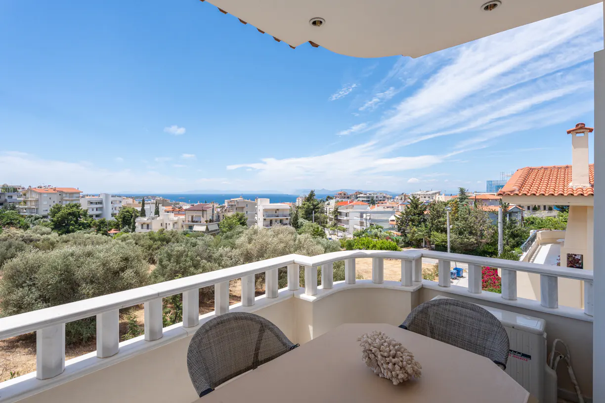 Balcony with a table and chairs overlooking a coastal town, olive trees, and the blue sea under a clear sky.
