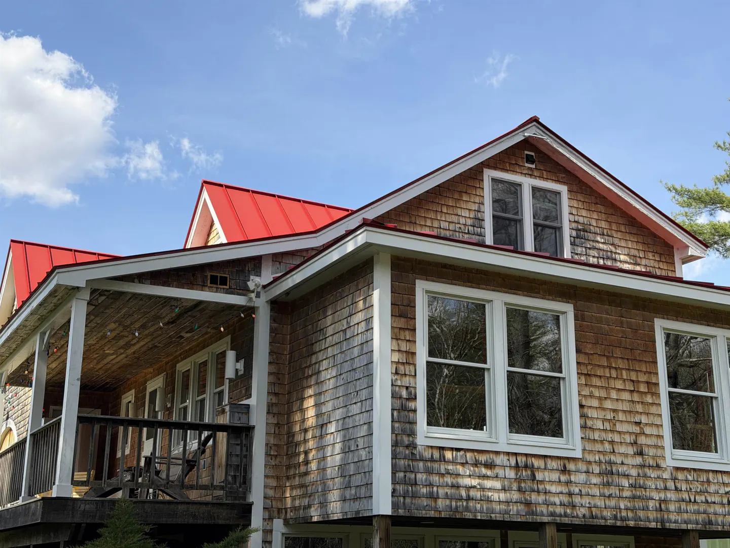 Exterior view of a house with cedar shingles and a bright red metal roof under a blue sky. A covered porch with white pillars is on the left.