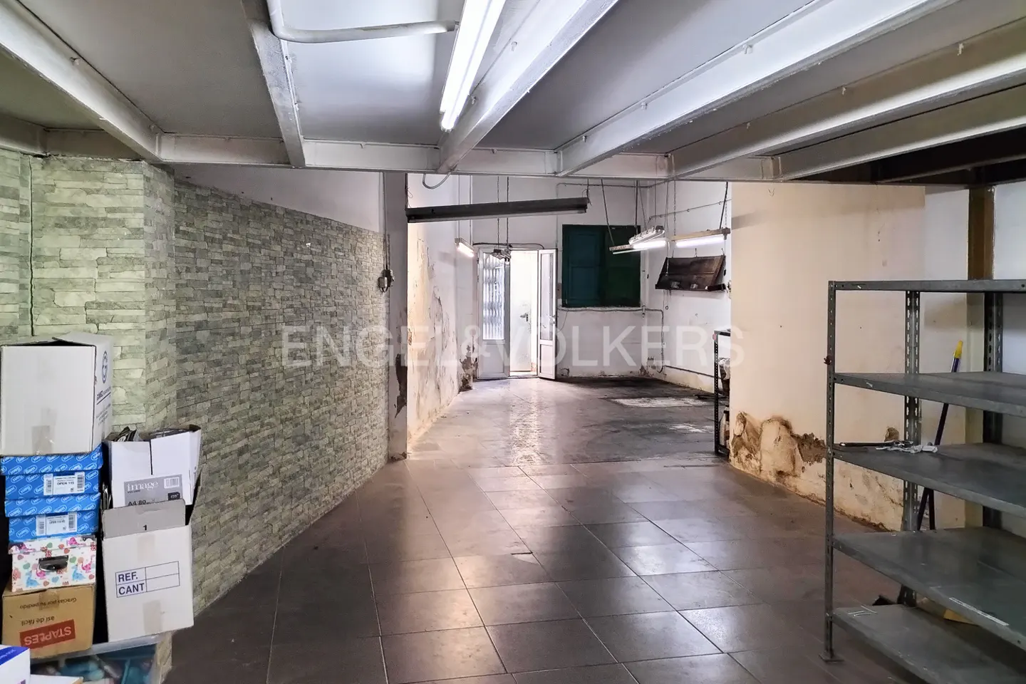 A commercial space with gray tile floors, exposed brick, and metal shelving. Boxes are stacked near the wall.